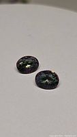Photo showing two oval cut mystic topaz gemstones on a white surface, highlighting their iridescent color and faceted surface.