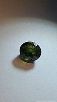 Oval cut green-yellow sapphire gemstone positioned on a plain surface, showing dark green color with internal inclusions.