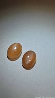 Pair of oval peach moonstone cabochon gemstones on a neutral background showing size and color, highlighting smooth polished surface and subtle shine.