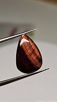 Close-up image of a pear-shaped red tiger's eye cabochon gemstone held with tweezers showing polished surface and light reflection.