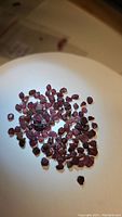 Photo showing a collection of small natural rough garnet stones scattered on a white dish under lighting, revealing their red to purple coloration and rough irregular shapes.