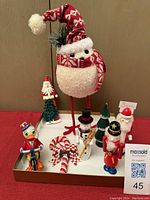 Front view of assorted holiday decorations including bird with knit hat and scarf, duck on motorcycle, Santa with tree, candy cane, and four place card holders arranged on white tray.