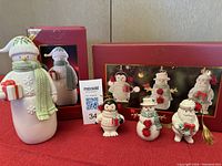 Front view photo of all figurines and ornaments including Chilly Cheer snowman, Merry Little Christmas set of three ornaments, and Yuletide figurine ornaments in front of their red boxes on red cloth background.