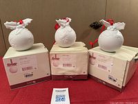 Three matte white NAO by Lladro holiday ball ornaments displayed in original boxes against a brown background with red surface.