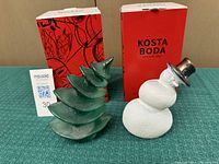 Two figurines side by side, tree in green textured glass with stylized layers, and snowman in white textured glass with black metallic hat, with red Kosta Boda boxes behind.
