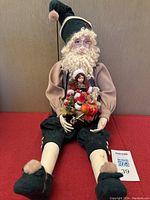 Full view of Kris Kringle sitting figurine on red surface with holiday toy collection in lap, showing green pants and slippers.