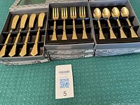 Three open boxes displaying 12 gold electroplate butter spreaders, 12 cocktail forks, and 12 demitasse spoons, all arranged neatly in black packaging.