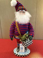 Front view of MacKenzie Child styled Santa in red and blue striped sweater and hat, holding black and white checked mailbag with holly and letters, standing on icy base with checkered border.
