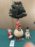 Santa on Snowball Tree with two snowpeople figurines in front on green cloth surface, visible 16 ornaments on tree