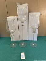 Three handmade clear glass candle holders of varying heights standing on a green mat with white boxes behind them.
