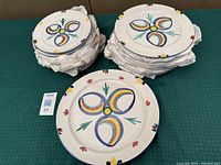 Top view of stacks of colorful Studio Nova Carnivale earthenware plates including salad and dinner plates, and a serving platter on a green surface.