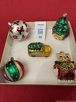 Five different glass ornaments on a white tray shown together: poinsettia ball, owl, yellow train car, green striped ball, and Christmas tree train car.