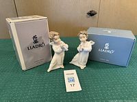Two porcelain angels playing flutes with their original Lladró branded boxes behind them on a green textured surface.