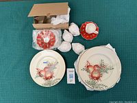 Photo showing Villeroy & Boch Toy's Delight demitasse cups wrapped in paper and red scalloped saucers with holiday decoration alongside Lenox cream-colored plates with apple and berry design.