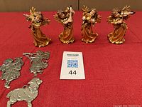 Four wooden golden angel musician figurines playing violin, lyre, flute, and trumpet, posed standing on red fabric background alongside three silver pewter ornaments (two angels and a sheep).