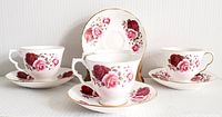 Photo showing four teacup and saucer sets arranged with some cups placed on saucers and one cup centered in front with a saucer to the side, all featuring pink and red rose floral patterns with gold trim.