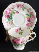 Close-up of the tea cup sitting in front of the saucer showing the rose floral pattern and gold trim along the cup rim, handle and base.