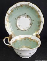 Photo showing teacup with handle and the saucer behind, displaying green color and gold decorative pattern.
