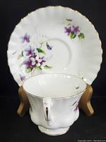 Front view of the white bone china saucer with purple violet and green leaf floral pattern and gold trim. The teacup is positioned in front showing the floral pattern on the side and ornate handle.