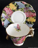 Front angle showing teacup with floral pattern and gold rim in front of matching saucer with colorful flowers on black background.