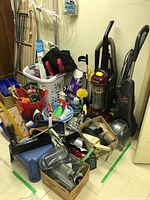 Wide view of assorted cleaning items including upright vacuums, vacuum parts, cleaning sprays, ironing board, and laundry basket.