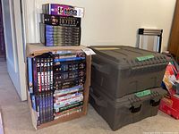 Overview showing two gray plastic storage bins and a cardboard box filled with DVD sets including The Good Guys and Arthur Hailey's The Hotel on top