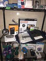 Photo of Sylvania 10.1" portable DVD player box, Boostmi jump starter box, Midland weather alert radio, Philips recording accessories, and various small electronics items on wire rack shelf.