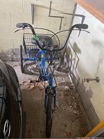 Front view of blue adult tricycle parked in a garage area between a car and a wall. Shows handlebars, front wheel, and rear basket.
