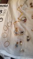 Overview of various jewelry items laid on a white cloth, including gold-tone bracelet, hoop earrings, pink stone ring, silver necklace, and multiple sets of earrings.