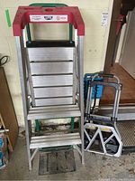 Full view of the aluminum step ladder, the luggage cart, and collapsible seat arranged near a garage wall.