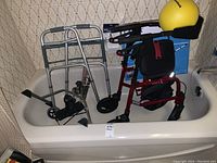 Bathtub holding two walkers (one silver standard walker, one red rollator walker), a bed support rail, and a small yellow bike pedal