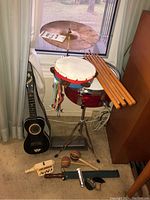 Overall view of the lot showing snare drum, cymbal, tambourine with ribbons, drumsticks on wooden furniture, maracas, recorders, and stringed instrument leaning against wall.