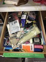 Drawer packed with various household and utility tools including screwdriver set, cable ties, cutters, adhesive products, and packing materials.