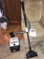 Front and side view of vacuum cleaner showing hose, wand, and power nozzle on carpet inside a living room.