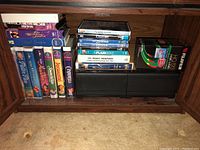 Shelf view showing row of Disney and feature film VHS tapes at left, DVD stack and promotional tape in center, blank Fujifilm VHS tape boxes at right