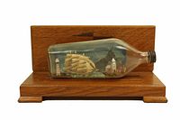 Ship in a bottle with multi-mast sailing ship inside, lighthouse and coastal decorations in background, set in a glass bottle on a polished wooden base.