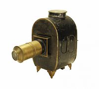 Side view of the black metal magic lantern featuring a brass lens and ornamental legs, showing wear and scratches.