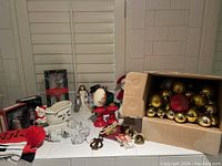 Photo showing a variety of used Christmas decorations on a white tiled surface, including a box of ornaments, figurines of Santa, angel, snowman, candle holders, and glass items.