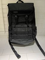 Front view of black Timbuk2 backpack showing flap with two buckle closures, front zippered pocket, and mesh side pocket.