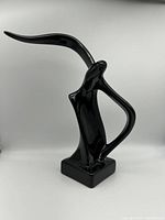Single black abstract bird sculpture with high arched beak on square base photographed from front.