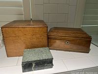 Three boxes placed on a white tiled tabletop including a large wooden box, medium wooden inlaid box and a small greenish stone box