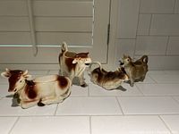 Four ceramic cow creamers in brown and white glaze arranged on a white tiled surface.