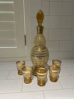 Photo of the gold-striped glass decanter with stopper and six matching glasses on a white tiled surface.