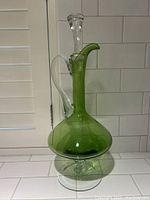 Full view of green glass decanter showing bulbous base, clear handle, and stopper on white tiled surface.