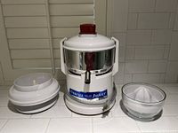 Full view of the Omega juicer main unit, featuring stainless steel body and white plastic lid, with separate citrus reamer and glass bowl placed beside.