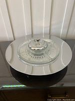 Front angle photo showing the glass Lazy Susan on a reflective surface, highlighting the segmented clear glass tray and chrome base with handle.