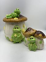 Front view of the ceramic mushroom canisters with the green frog figurines placed on and around them.