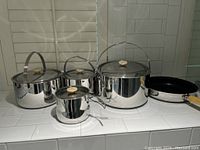 Front view of all items showing 3 stainless steel covered pots and 1 frying pan with wooden handle knob on a white tiled counter.