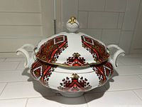 Porcelain tureen with lid displayed on a white tiled surface, showing side view with handles and ornate lid knob.