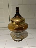 Front view of vintage amber glass Empoli jar with gold decorative accents and rounded finial lid, on clear pedestal base.